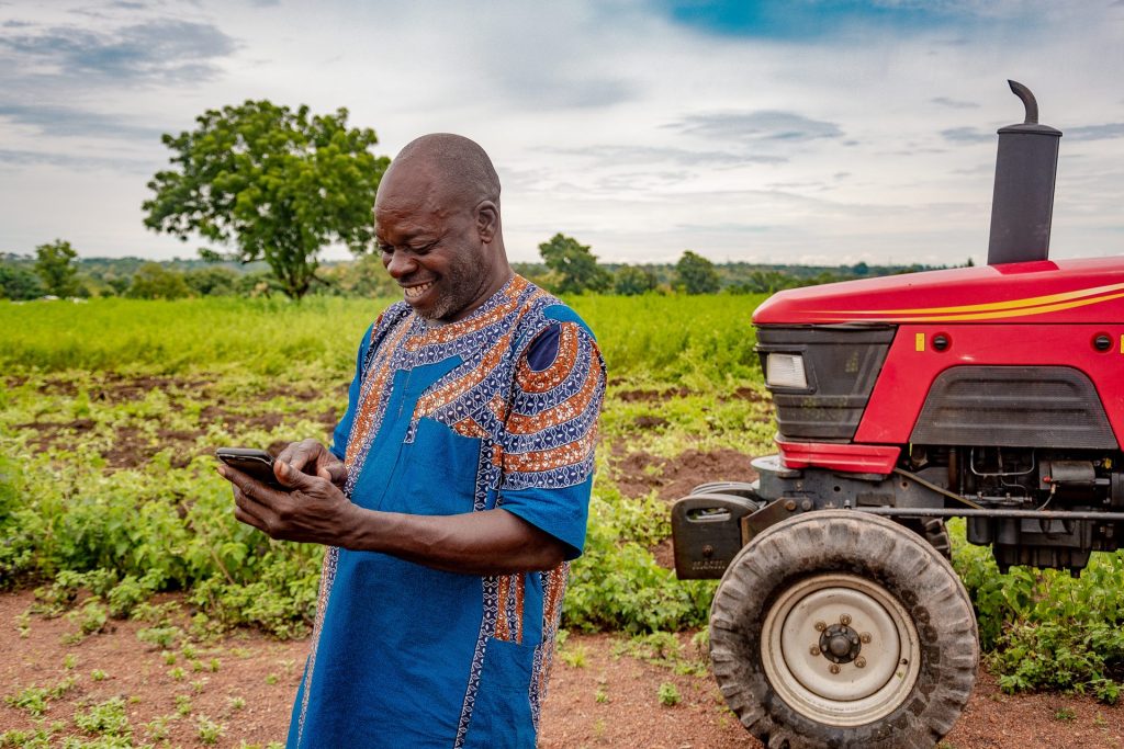 A farmer standing next to a tractor on the field is looking at his mobile phone smiling.
